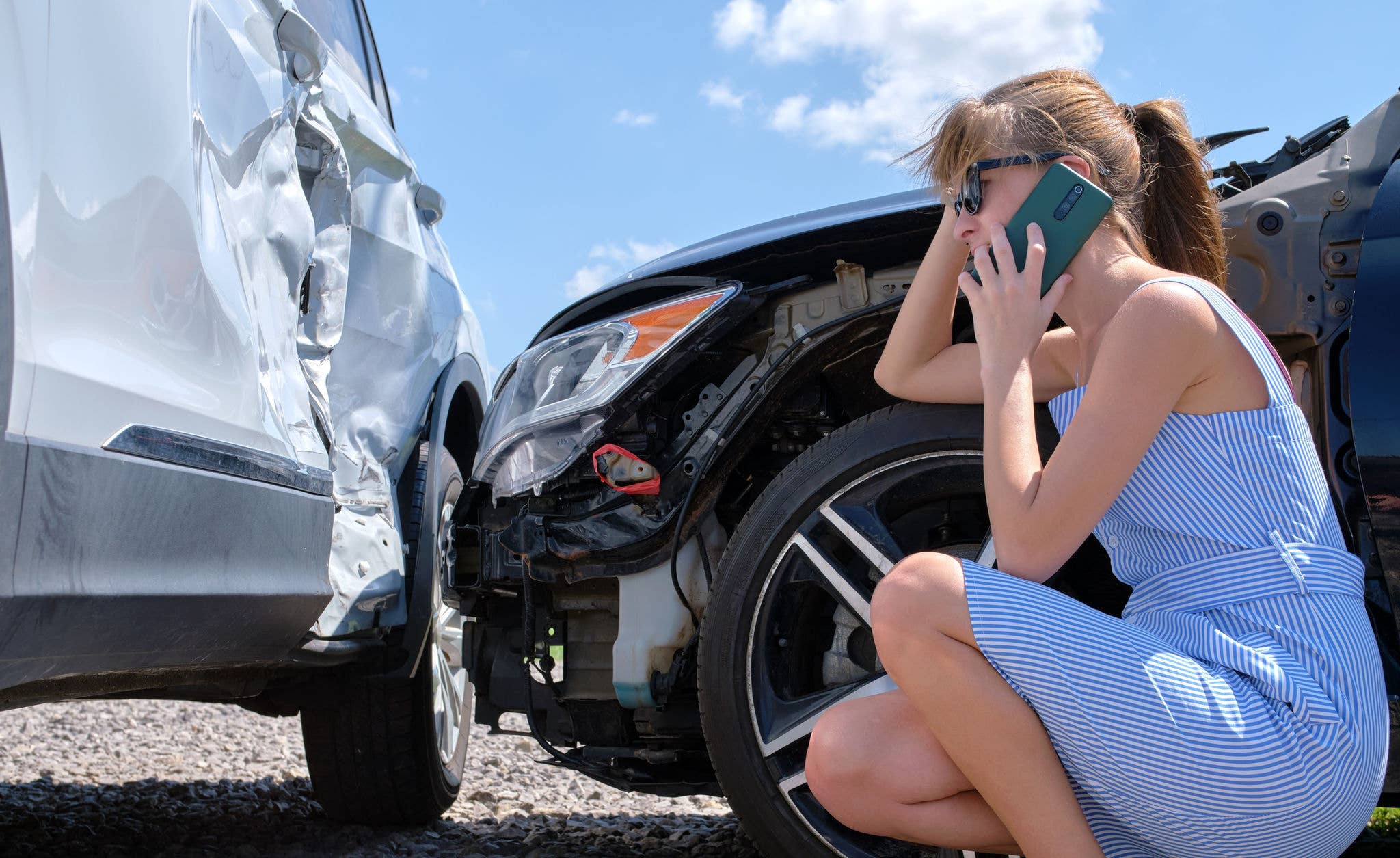 A young woman on the phone inspecting damage to her car after an accident.