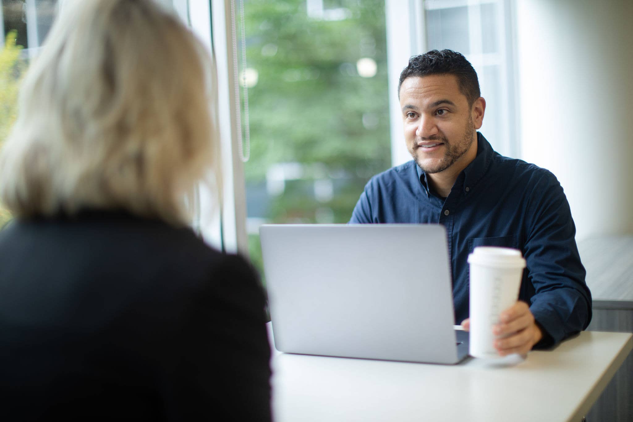 Man holding a coffee cup behind a laptop while talking to a woman.