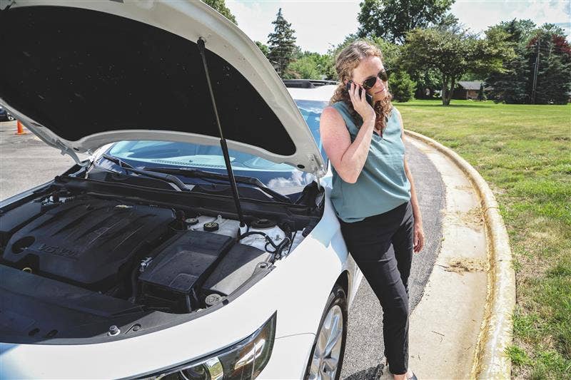 Woman stands with her phone next to her broken-down car.