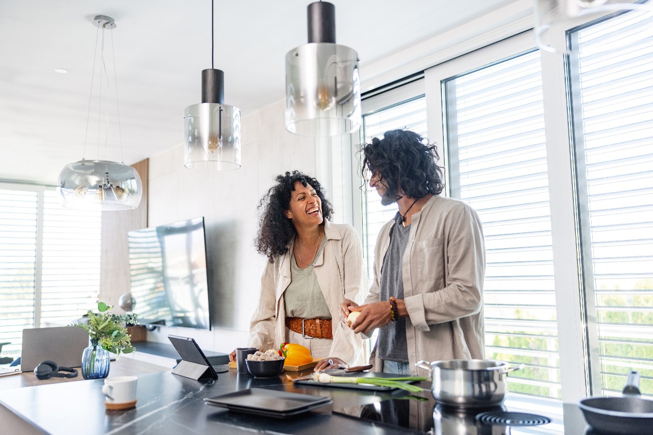 Multiracial mid-adult couple in their early 30's smiling to each other while cooking together in the kitchen and following a recipe in the digital tablet surrounded with different ingredients and kitchen accessories on a sunny morning.