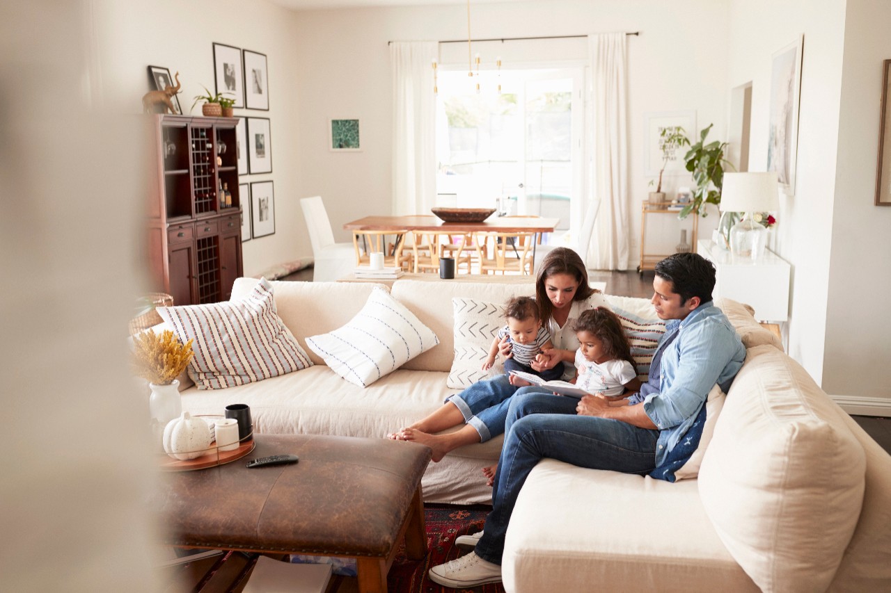Young Hispanic family sitting on sofa reading a book together in the living room, seen from doorway