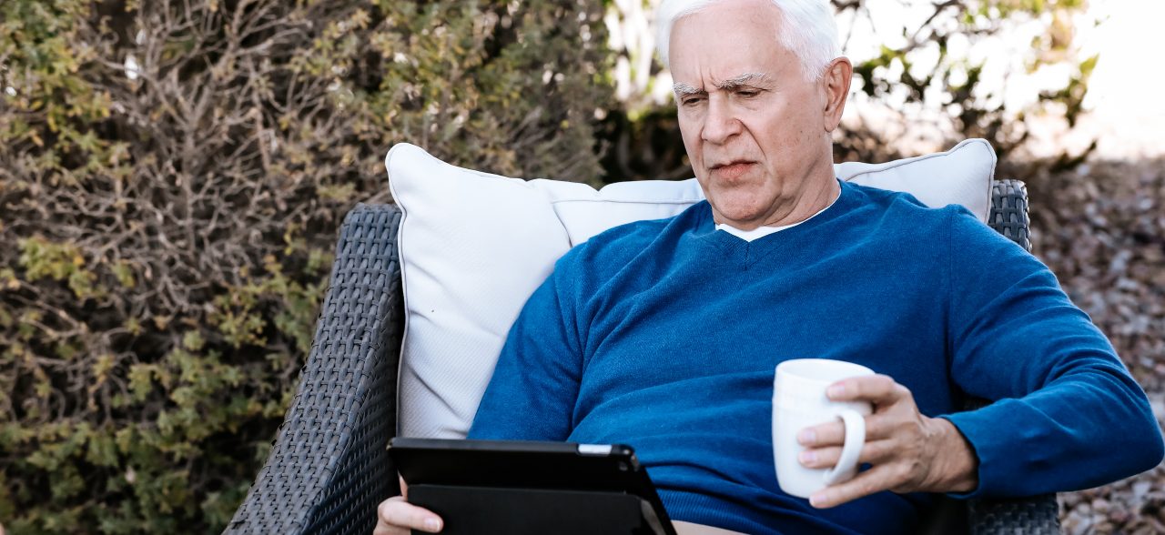 Senior man sitting on patio furniture drinking coffee and looking at tablet